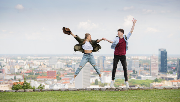 A cheerful young couple travelers in city on holiday, jumping. Cityscape in the background.