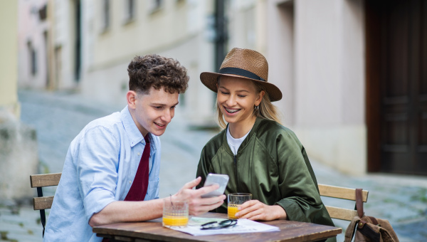 Happy young couple travelers using smartphone in city on holiday, sitting in outdoor cafe.