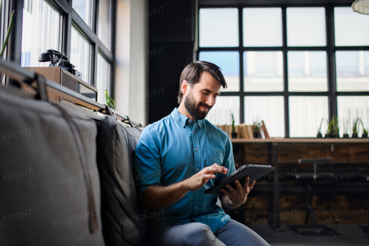 Portrait of young businessman with tablet sitting indoors in office, looking at camera.