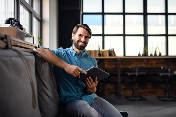 Portrait of young businessman with tablet sitting indoors in office, looking at camera.