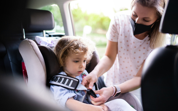 Mother putting small toddler daughter in car seat before trip, wearing face masks.