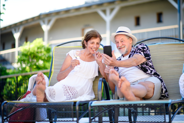 Front view of senior couple with wine and smartphone outdoors on holiday, relaxing.