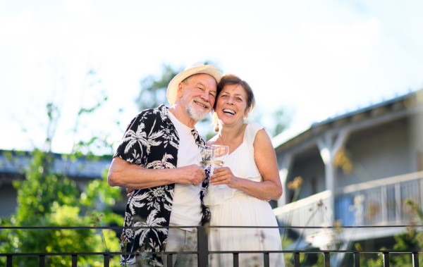 A senior couple holding wine outdoors on holiday, looking at camera.