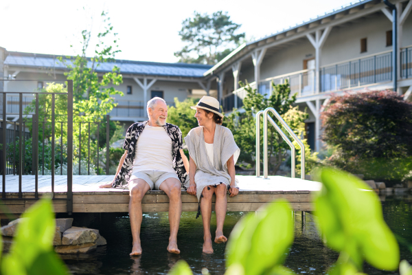 Senior couple sitting and talking outdoors by lake hotel on holiday, having fun.