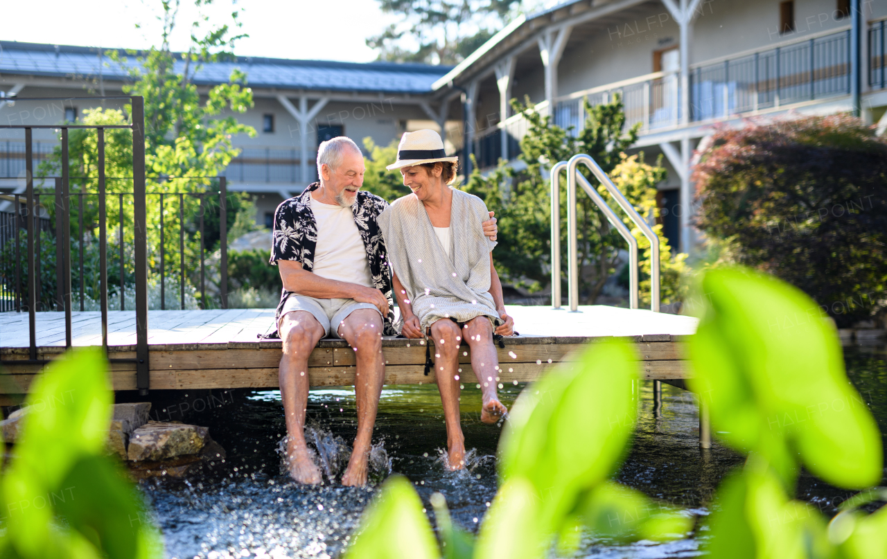 Senior couple sitting and talking outdoors by lake hotel on holiday, having fun.