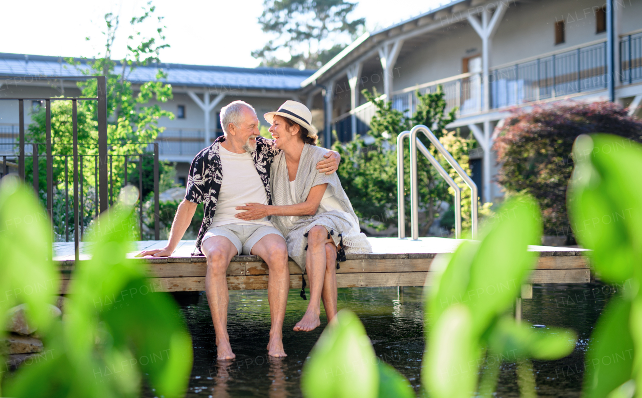 Senior couple sitting and talking outdoors by lake hotel on holiday, having fun.