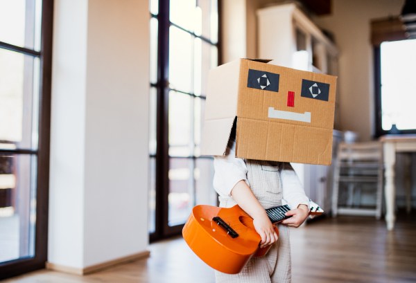 A toddler girl with carboard monster and guitar on head playing indoors at home.