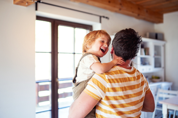 A mature father carrying toddler boy indoors at home, laughing.