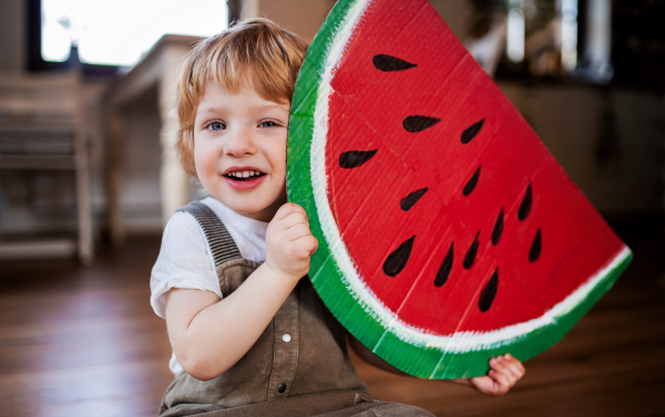 A toddler boy playing with large toy fruit indoors at home, eating fruit concept.