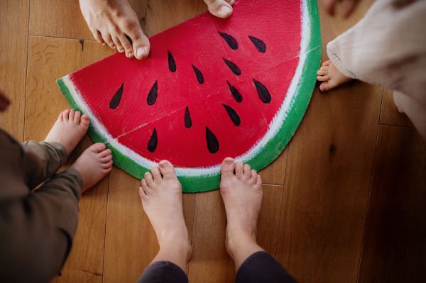 A top view of bare feet of family standing on large toy fruit indoors at home.