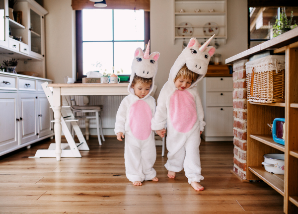 Two toddler children with white unicorn masks walking indoors at home.