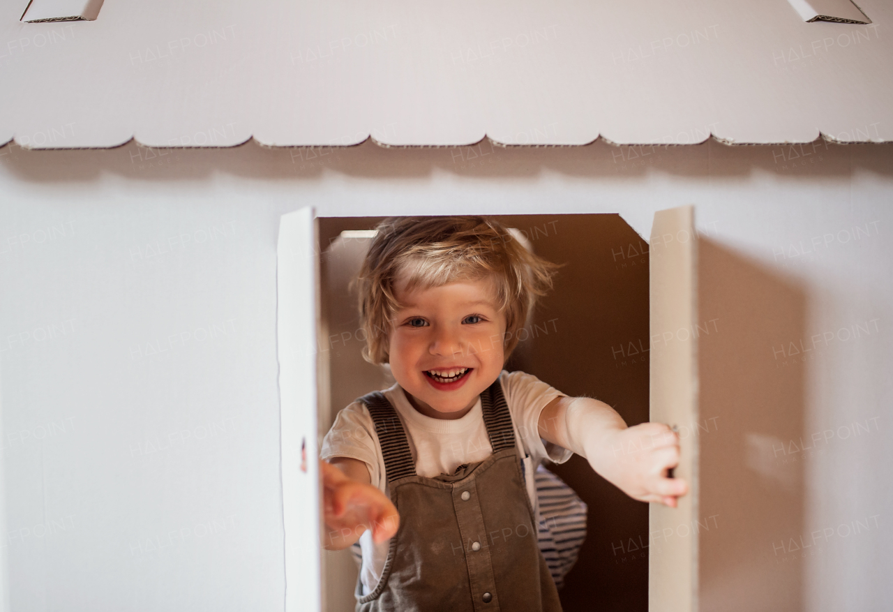 A toddler boy playing indoors with cardboard house at home, opening window.