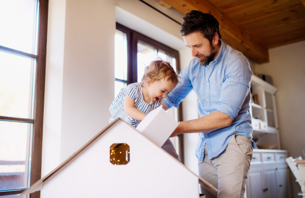 Mature father with small daughter playing with carboard house indoors at home.