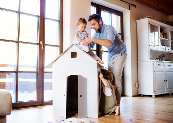 Two happy toddler children with father playing with paper house indoors at home.