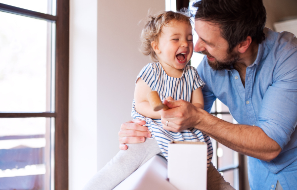 Mature father with small daughter playing with carboard house indoors at home.