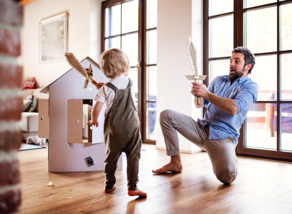 A toddler boy and father with carton swords playing indoors at home, fighting.