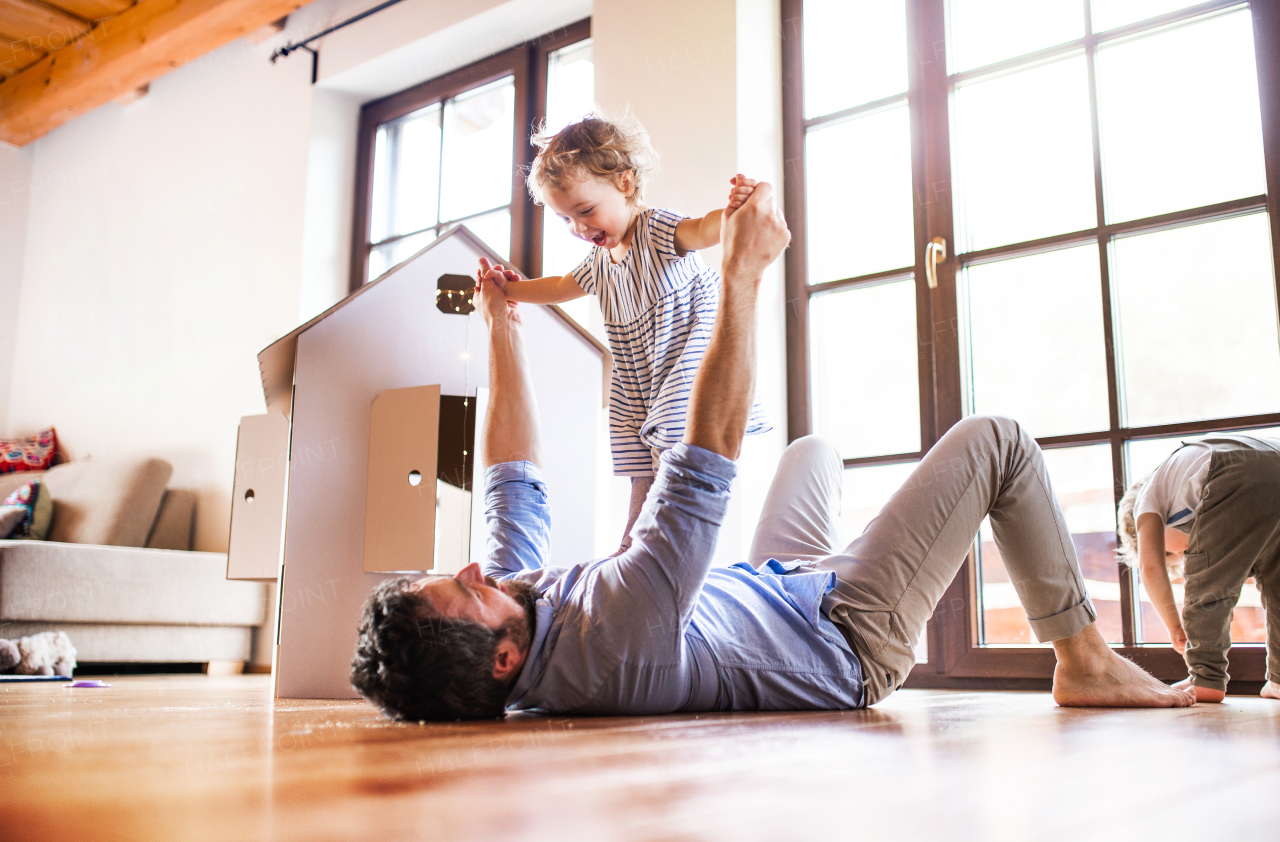 Two happy toddler children with a father and carboard house playing indoors at home.