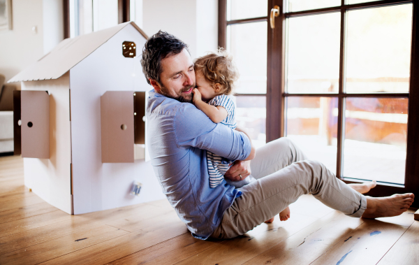 Two happy toddler children with a father and carboard house playing indoors at home.