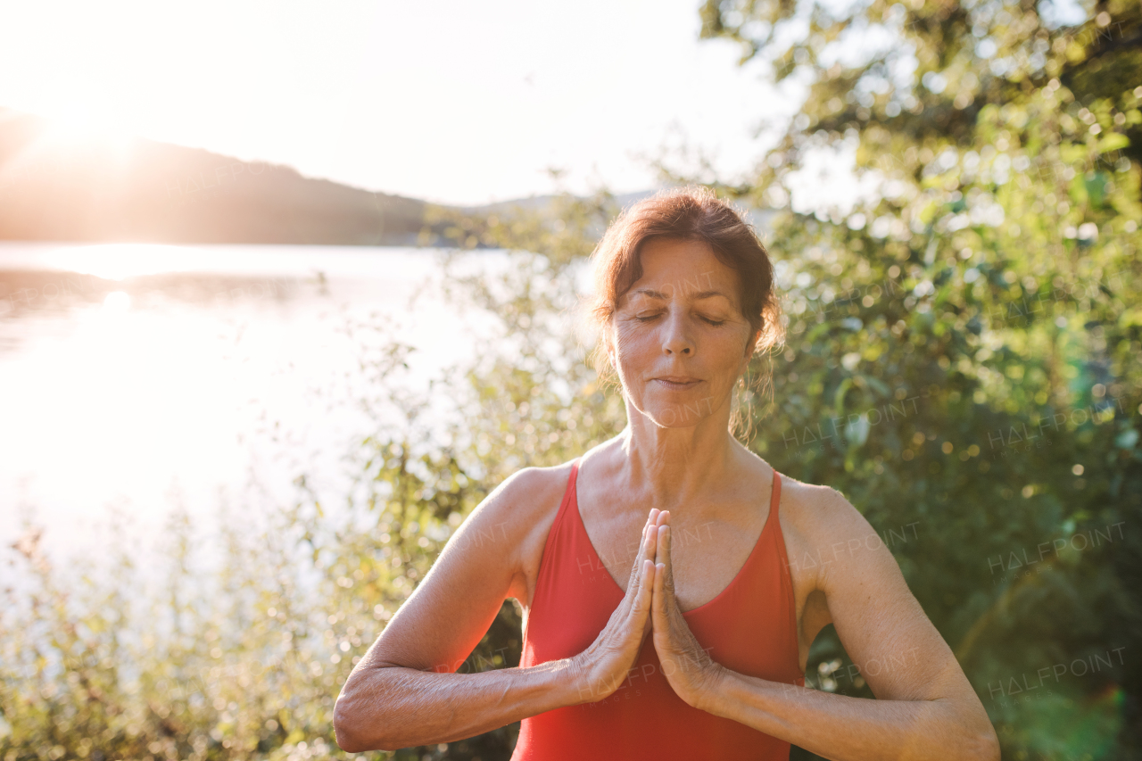 A senior woman in swimsuit standing by lake outdoors doing yoga exercise.