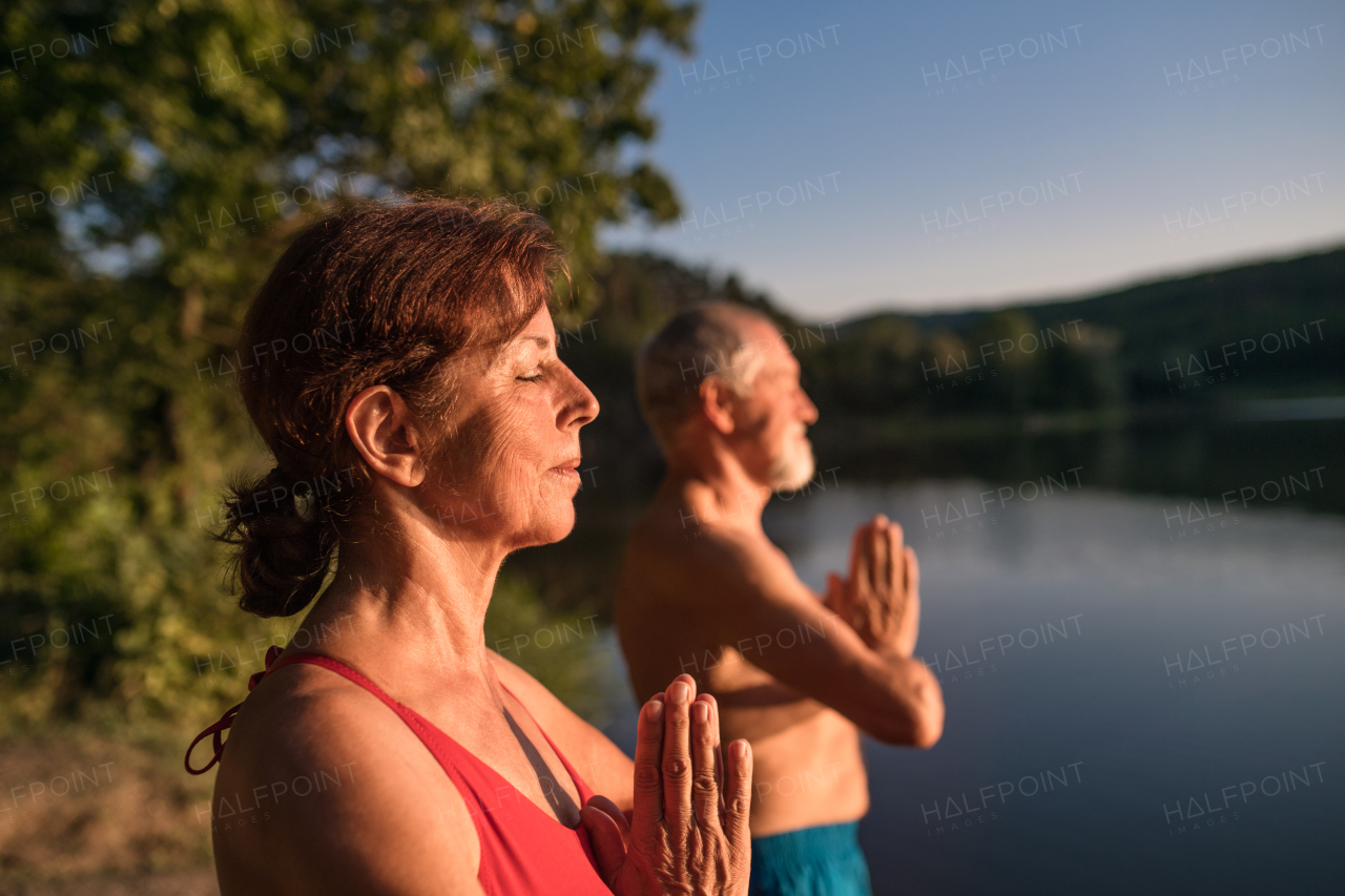 Senior couple in swimsuit standing by lake outdoors before swimming, doing yoga exercise.