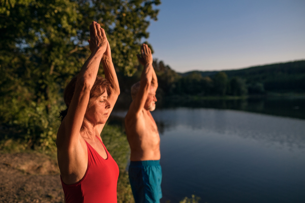 Senior couple in swimsuit standing by lake outdoors before swimming, doing yoga exercise.