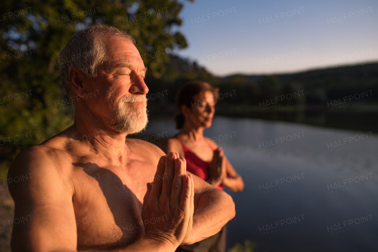 Senior couple in swimsuit standing by lake outdoors at dusk before swimming, doing yoga exercise.