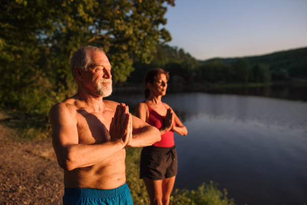 Senior couple in swimsuit standing by lake outdoors before swimming, doing yoga exercise.