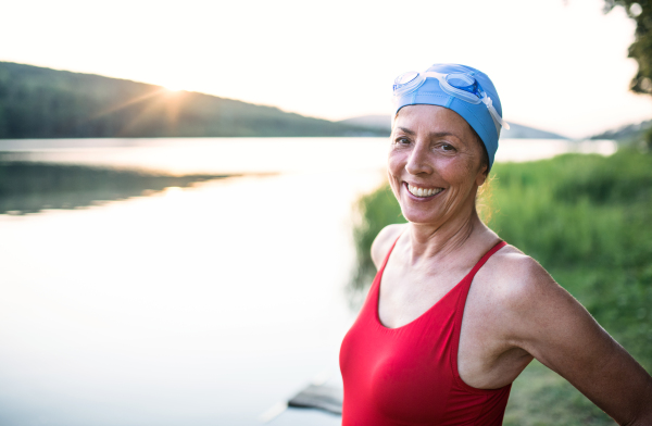 Cheerful senior woman in swimsuit standing by lake outdoors before swimming.