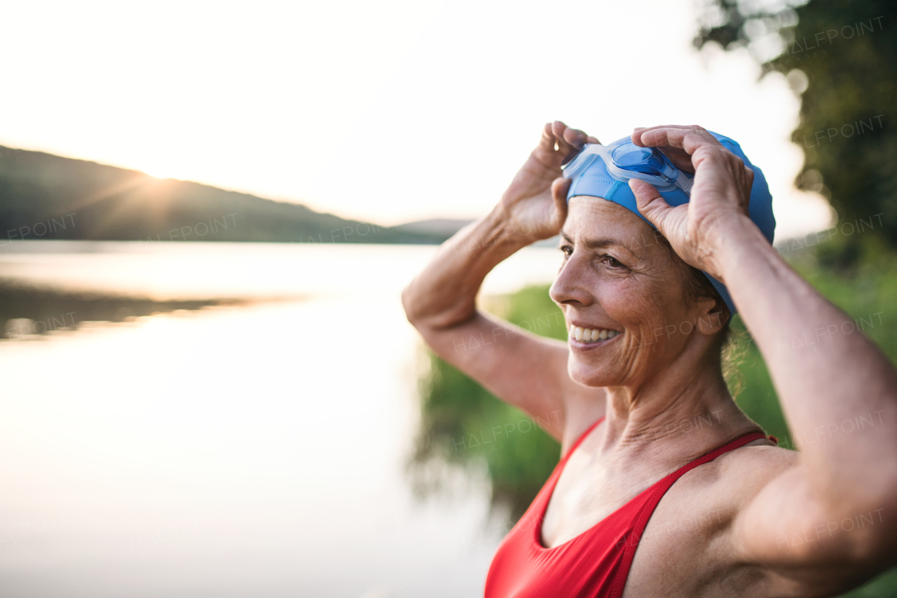 Cheerful senior woman in swimsuit standing by lake outdoors before swimming.