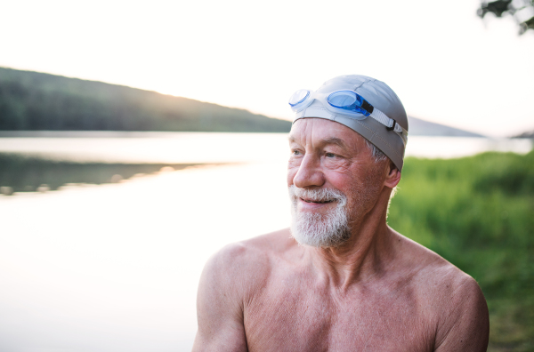 A senior man in swimsuit standing by lake outdoors before swimming.