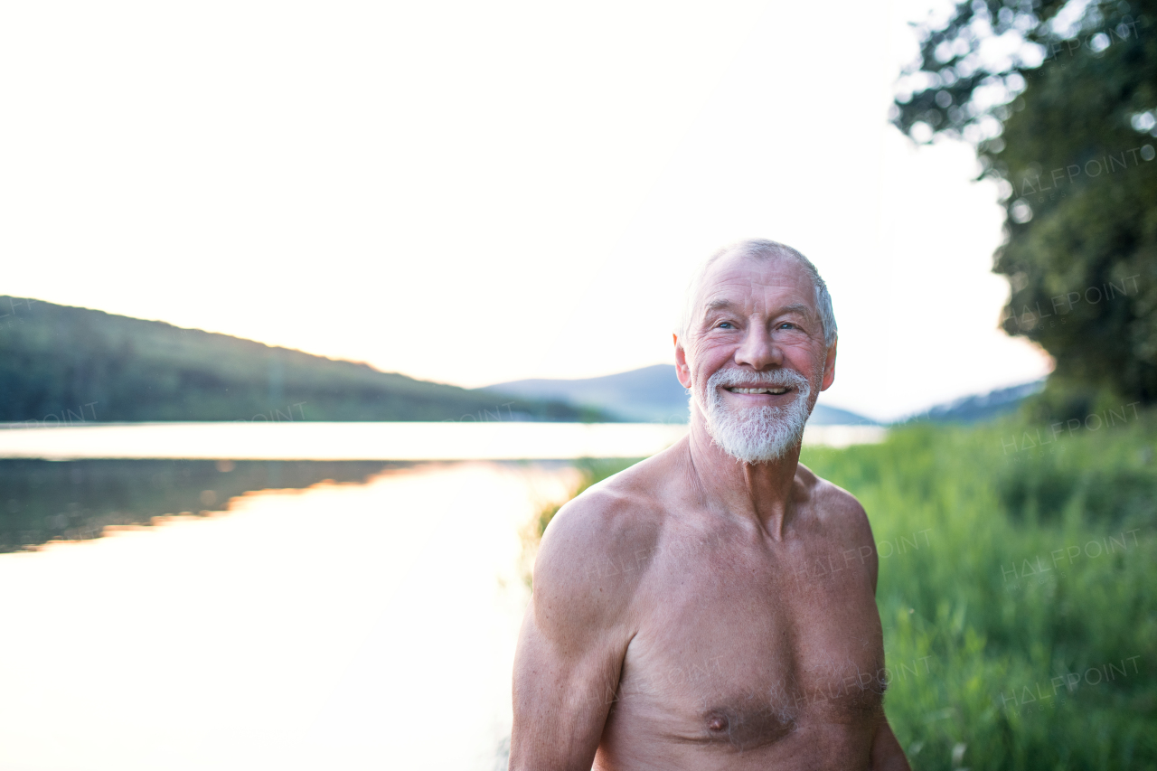 Senior man in swimsuit standing by lake outdoors before swimming. Copy space.