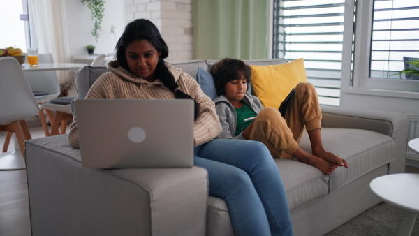 An Indian mother using laptop and her little son using tablet and sitting on sofa at home.