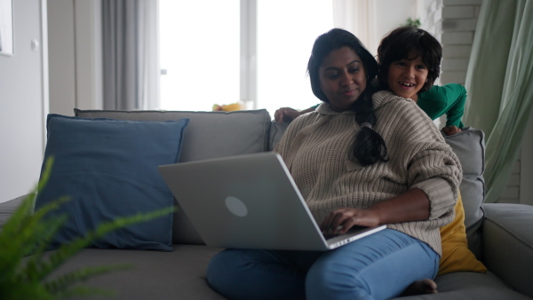 An Indian mother using laptop and her little son is suprising her on sofa at home.