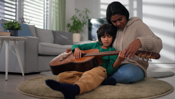 A little multiracial boy learning to play the guitar with his mother at home.