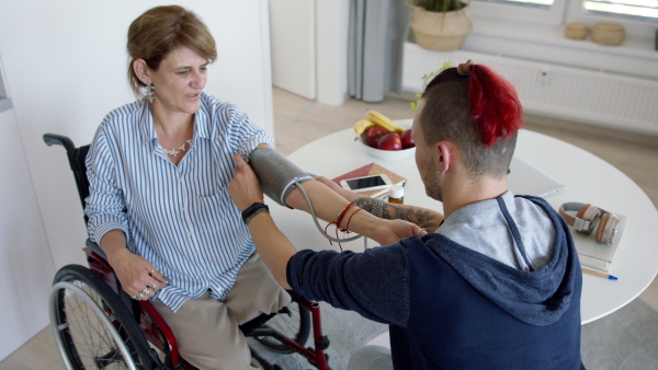 Two disabled people in wheelchair sitting at the table indoors at home, measuring blood pressure.