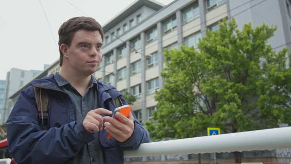 A young Down syndrome man using smartphone outdoors in city street, waiting for bus.