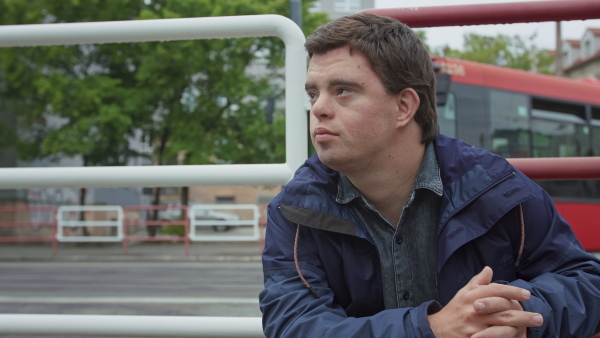 A close up of young Down syndrome man sitting outdoors in bus stop, waiting for bus.