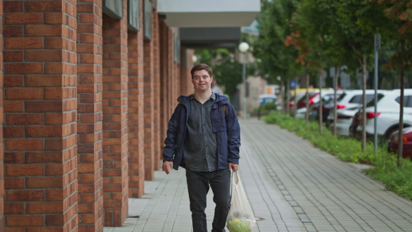 A man with Down syndrome walking outdoors in town street, holding mesh shopping bag and looking at camera.