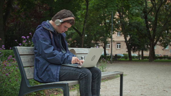 A man with Down syndrome with headphones using laptop and sitting on bench outdoors in town.