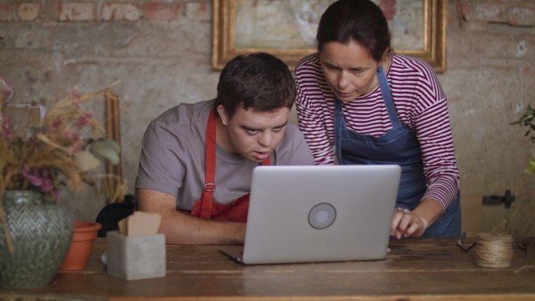 A young male florist with Down syndrome working on laptop with help of mentoring colleague indoors in flower shop.