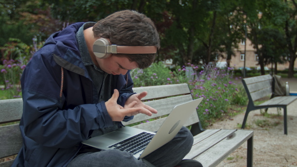 A man with Down syndrome with headphones using laptop and sitting on bench outdoors in town.