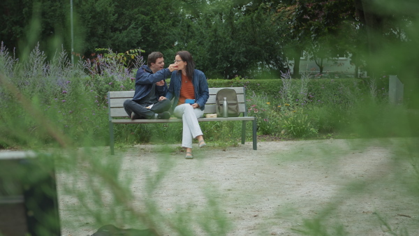 A happy mature mother with adult son with Down syndrome sitting on bench and feeding each other with apple outdoors in town.