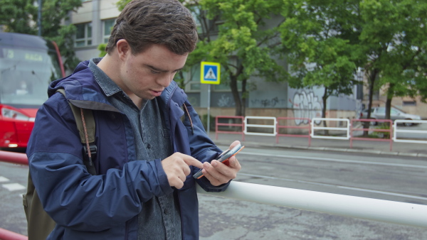 A young man with Down syndrome using smartphone outdoors in city street, waiting for a bus.