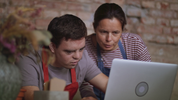 A young male florist with Down syndrome working on laptop with help of mentoring colleague indoors in flower shop.