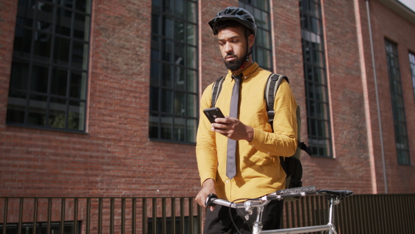 A young man commuter pushing bike and using smartphone in city.