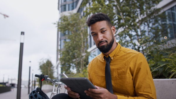 A young man commuter with bicycle sitting on bench using tablet and looking at camera in city.