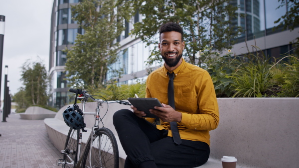 A young man commuter with bicycle sitting on bench using tablet and looking at camera in city.