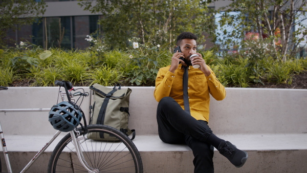 A young man commuter with bicycle sitting on bench and talking to phone in city.