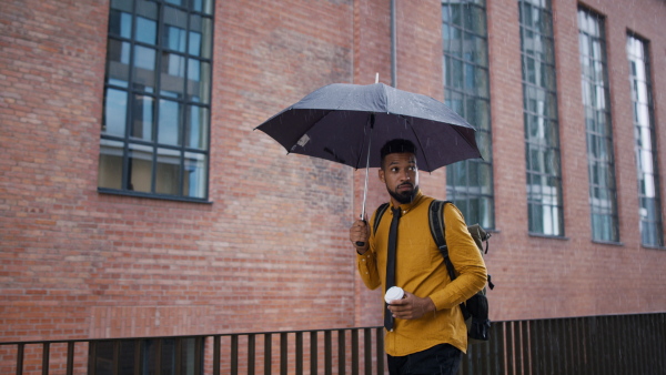 A young man commuter on the way to work with umbrella and takeaway coffee walking when raining.