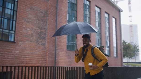 A young man commuter on the way to work with umbrella and takeaway coffee running when raining.